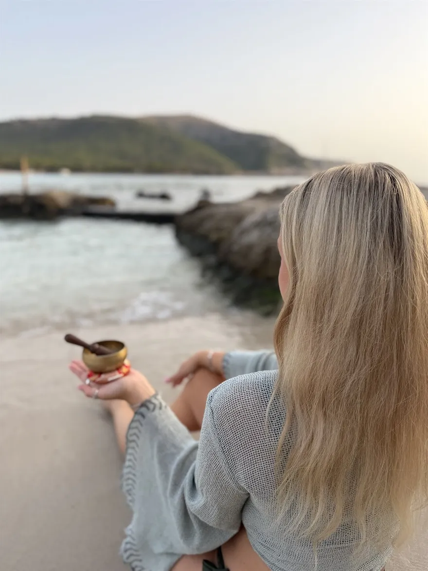 Nina Leismann sitzt mit einer Klangschale in den Händen am Wasser, von hinten fotografiert